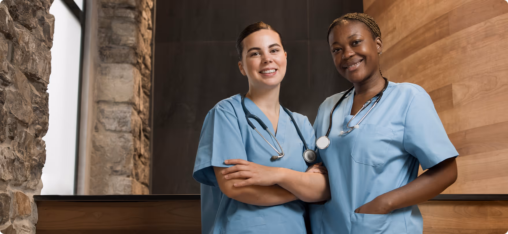 Two smiling healthcare professionals in blue scrubs with stethoscopes standing side by side indoors.