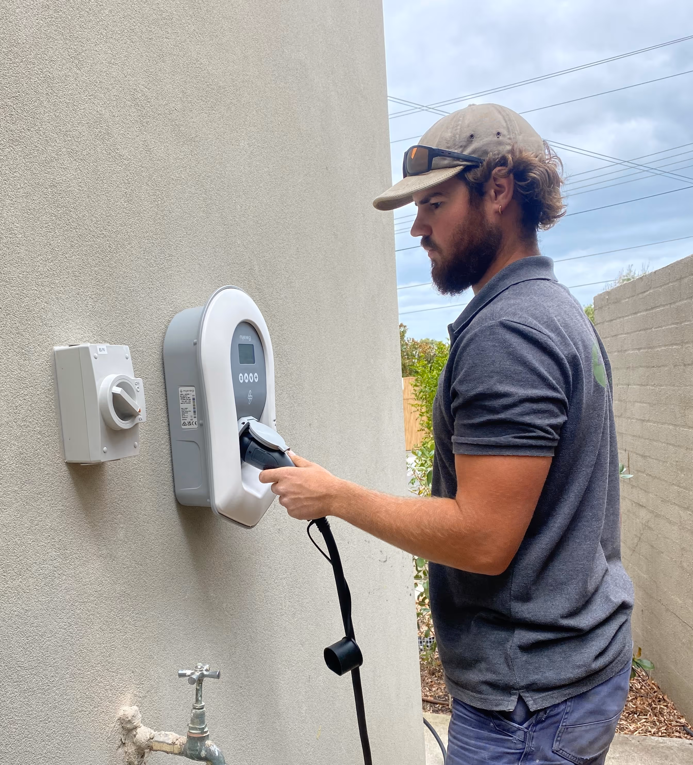 Man with beard and cap plugging an Zappi EV charger into a wall-mounted charging station.