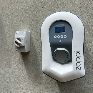 Wall-mounted Zappi electric vehicle charger with a digital display and control knob beside a separate rotary switch.