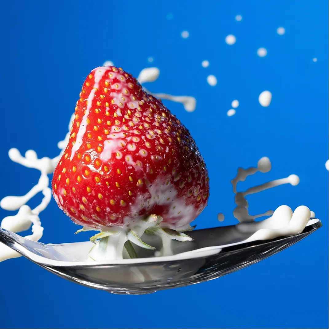 Close-up of a red strawberry with cream splashing on a metal spoon against a blue background.