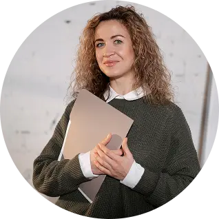Smiling woman with curly hair holding a closed laptop and wearing a dark sweater over a white collared shirt.