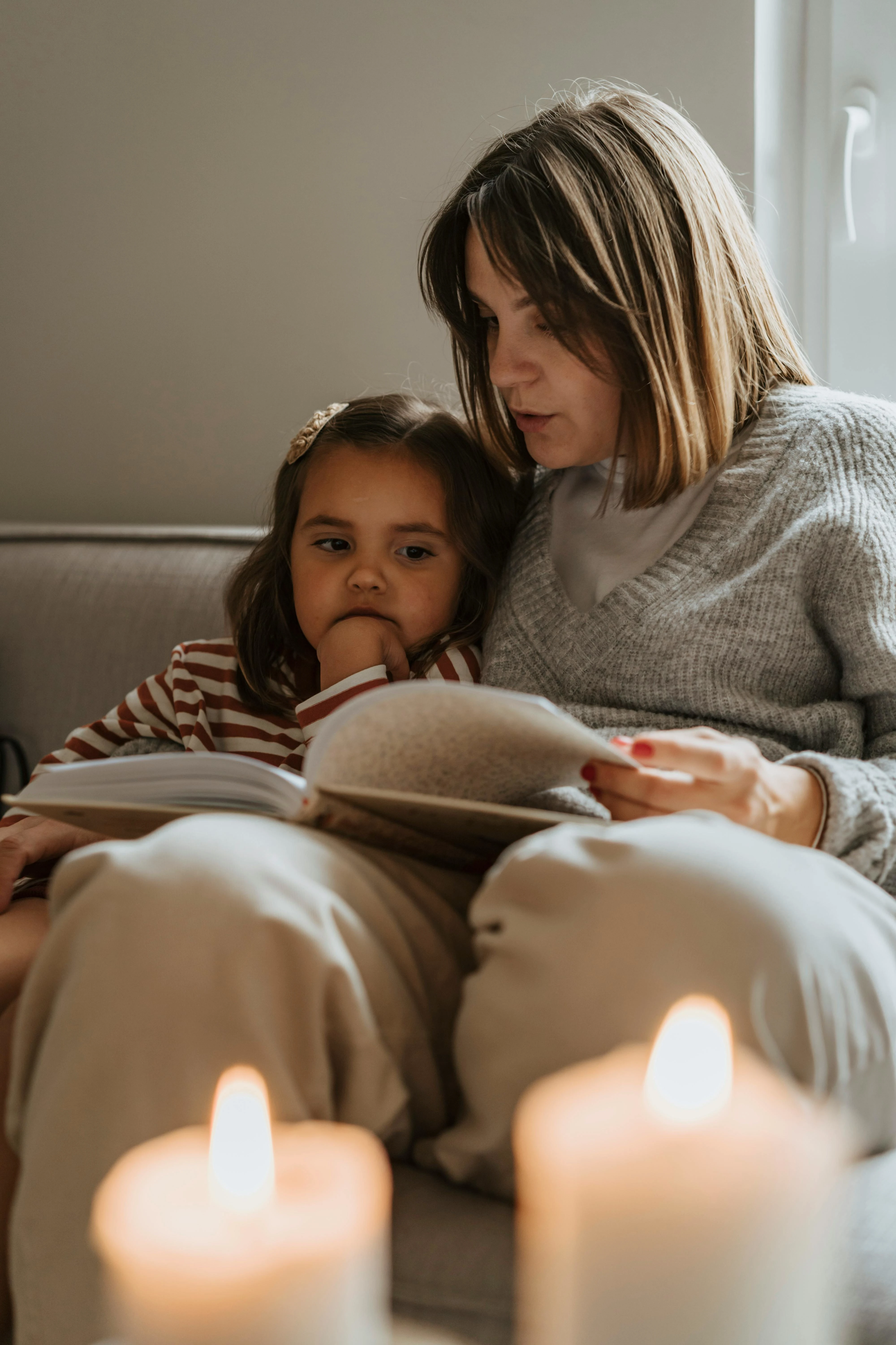 Kvinna läser bok för ett barn som sitter nära intill på en soffa med tända ljus i förgrunden.