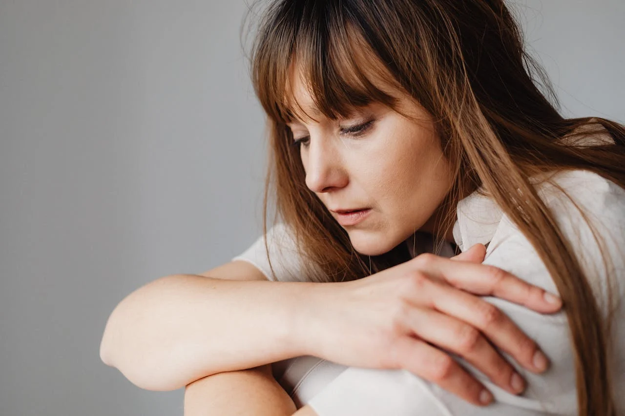 Närbild av en kvinna med långt brunt hår som ser nedåt med en sorgsen och eftertänksam blick, med armarna korsade runt knäna.