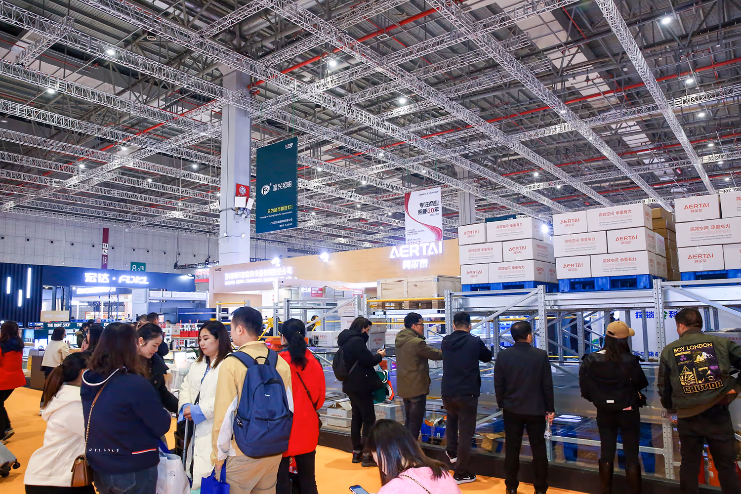 People gathered at an indoor trade show or exhibition with stacked boxes on shelves and booths in the background.