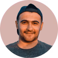 Headshot of a smiling man wearing a backward black baseball cap and a gray shirt against a soft pink circular background.