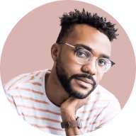 Man with curly hair, glasses, and a beard resting his chin on his hand against a soft pink background.
