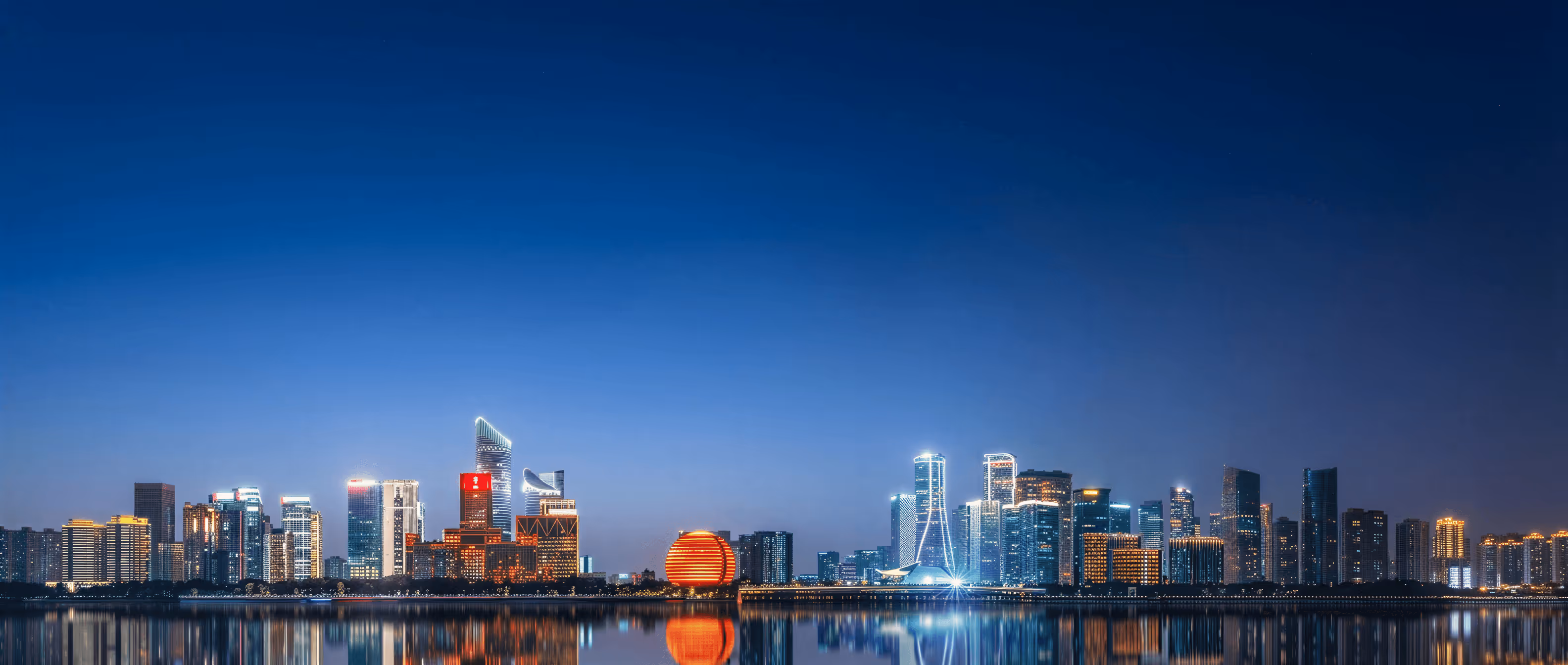 Panoramic night skyline of a city with illuminated modern skyscrapers reflecting on calm water.