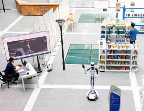 Interior of a modern space showing a person working on a laptop near a large screen, a delivery robot, and a man browsing items on store shelves.