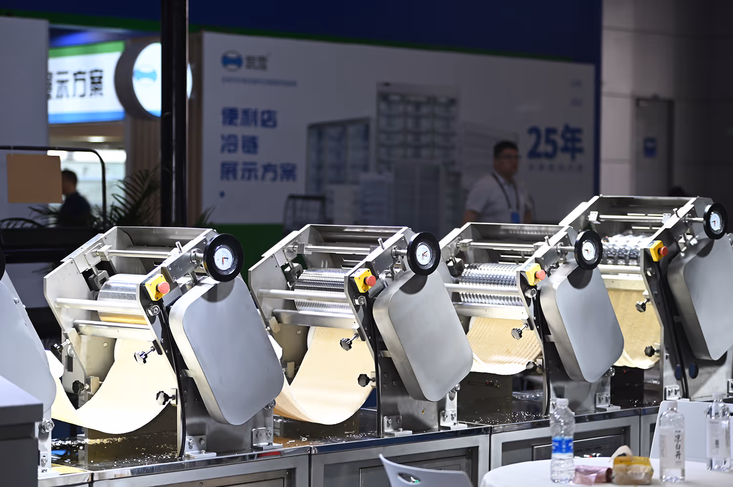 Row of industrial dough sheeting machines with rollers and dough sheets on a metal table in an exhibition setting.