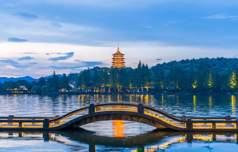 Curved pedestrian bridge over calm water with a lit pagoda and forested hillside at dusk.