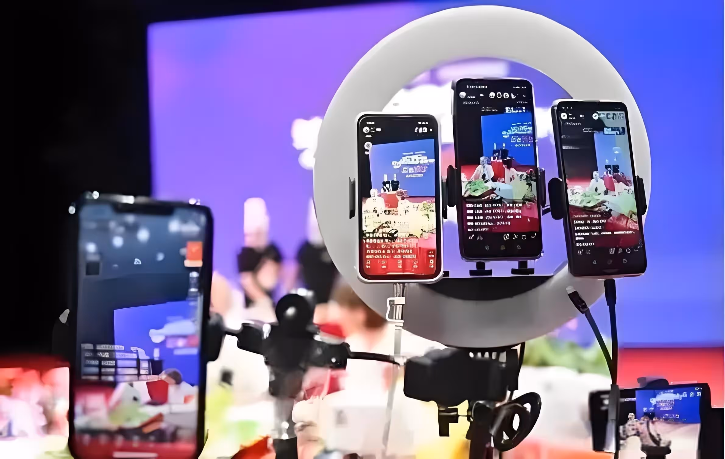 Multiple smartphones mounted on a tripod with a ring light, livestreaming a group of people seated at a table against a blue background.