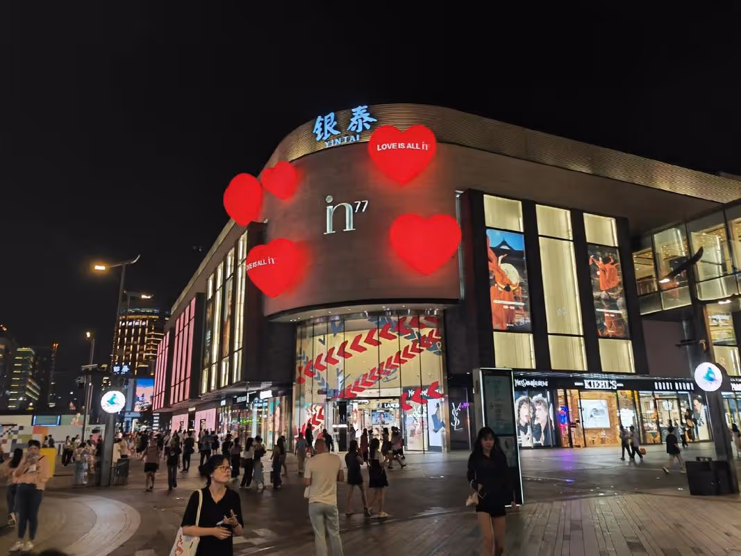 Night scene of a busy shopping mall entrance with illuminated signs and red heart-shaped decorations.