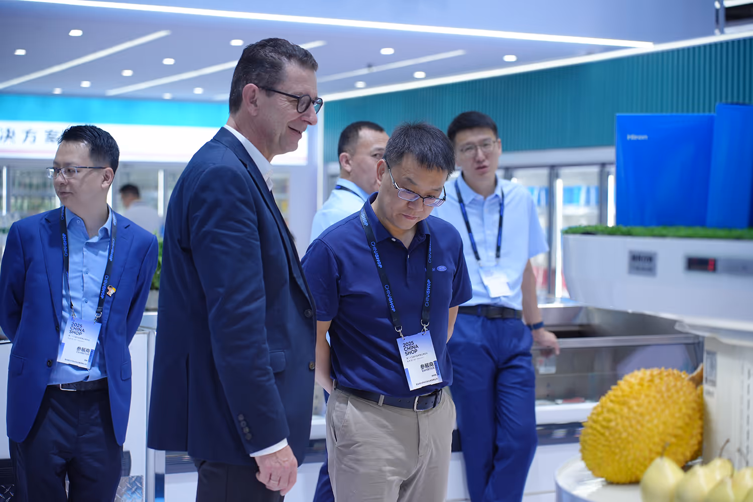 A group of five men at a trade show booth, two of whom wear badges labeled '2025 China Shop,' looking at a display with a large yellow spiky fruit and some pears.