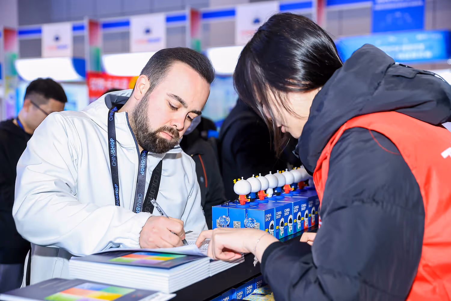 Man in a white jacket signing a document while a woman in a red vest assists him at a booth with blue products labeled China Shop.