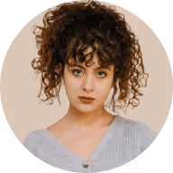Portrait of a young woman with curly hair wearing a light gray top against a beige background.