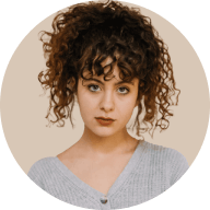 Portrait of a young woman with curly hair wearing a light gray top against a beige background.