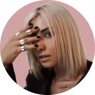 Blonde woman with short hair and black nail polish partially covering her face with her hand wearing multiple silver rings.
