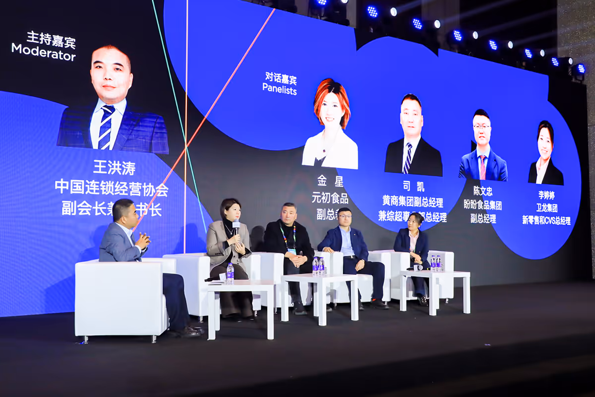 Panel discussion at a conference with five seated speakers on stage and a large screen behind showing photos and titles of the moderator and panelists.