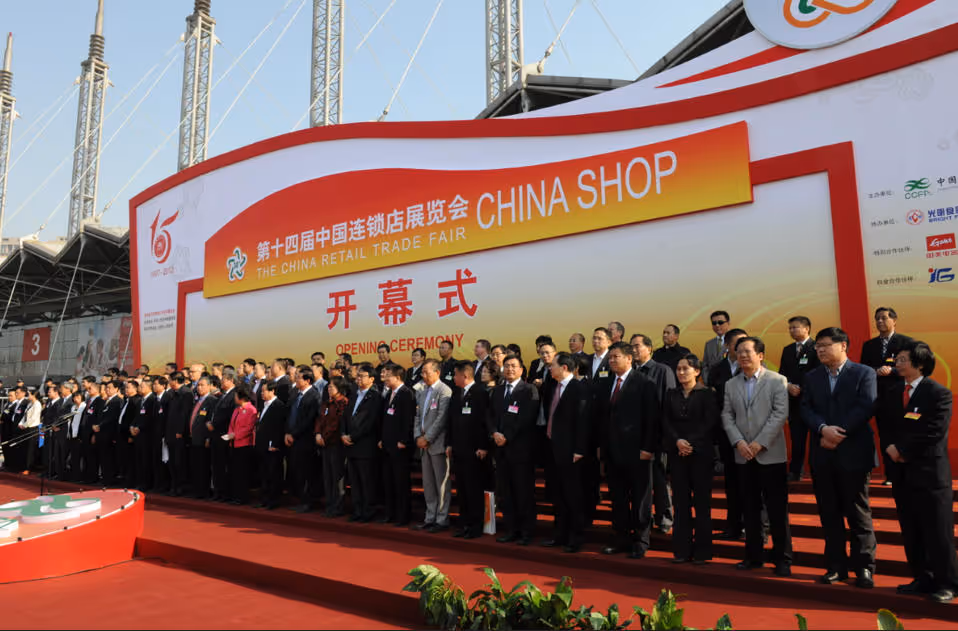 A large group of people dressed in formal attire standing on a red carpeted stage at the opening ceremony of The China Retail Trade Fair with a banner reading 'CHINA SHOP' in the background.