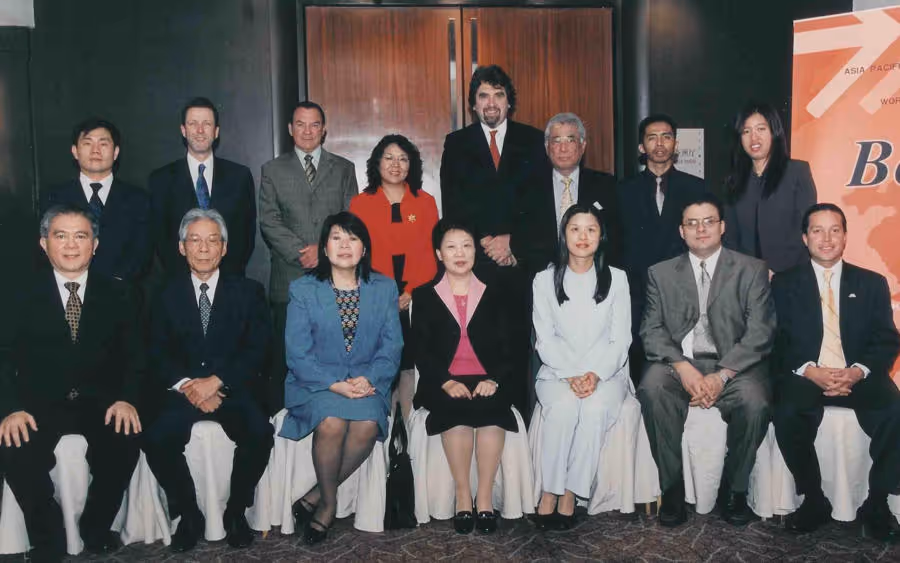 Group photo of fourteen people, men and women dressed in formal business attire, seated and standing in two rows inside a conference room.