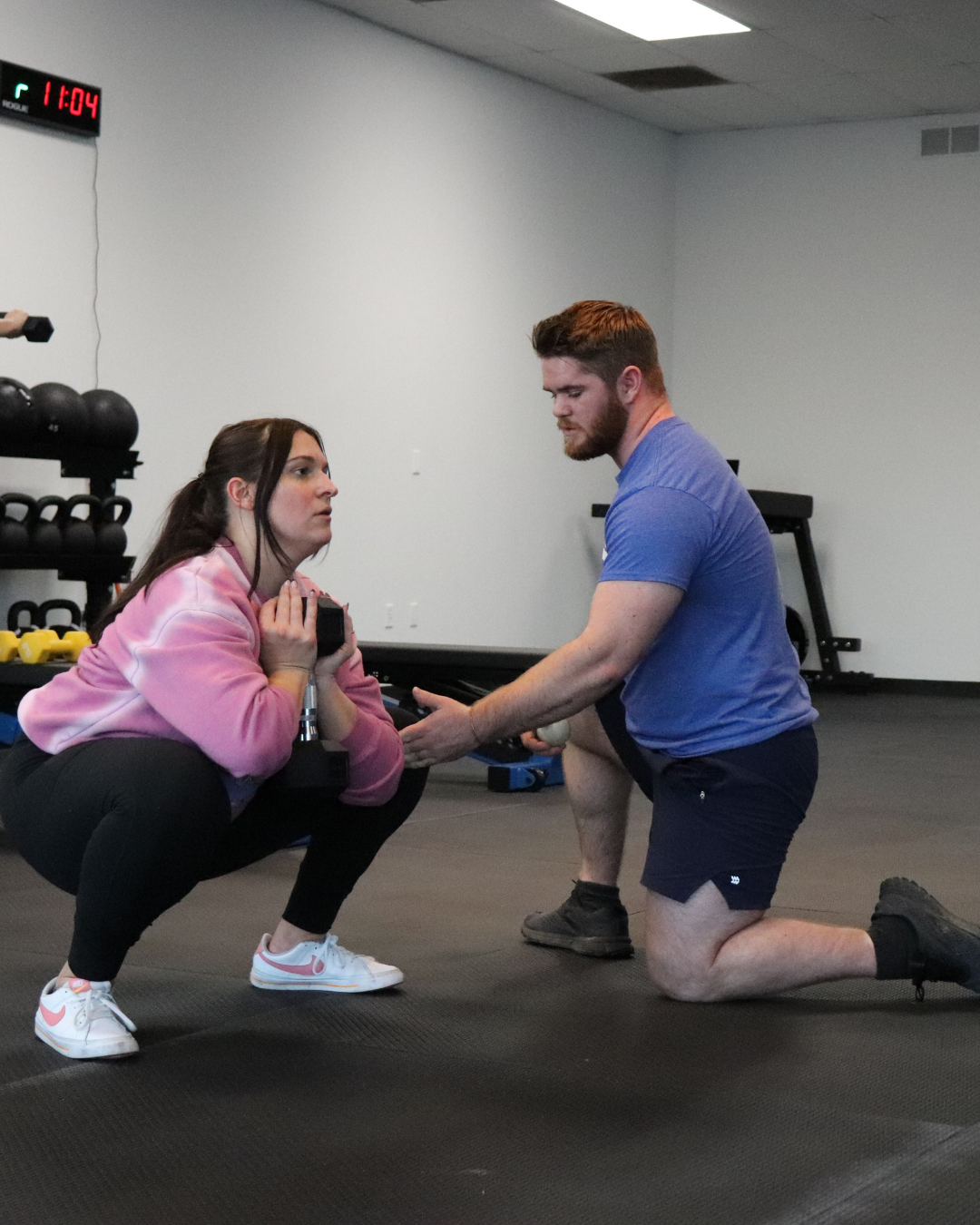 A woman in pink hoodie squats holding a dumbbell while a personal trainer kneels and coaches her in a gym.