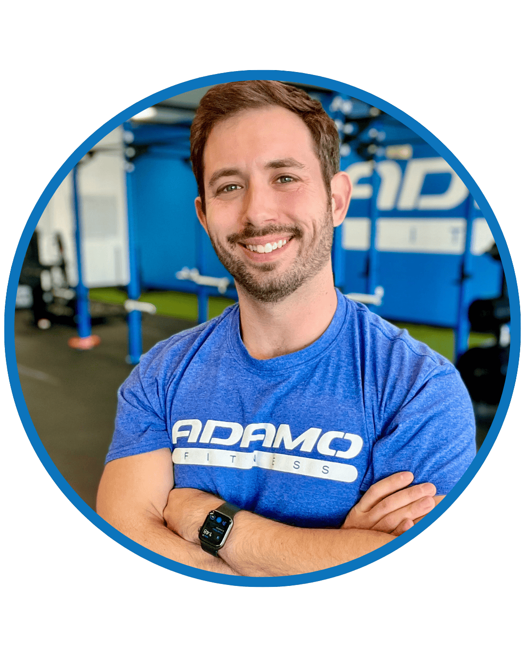 Smiling man in a blue Adamo Fitness t-shirt with arms crossed, wearing a smartwatch, inside a gym.