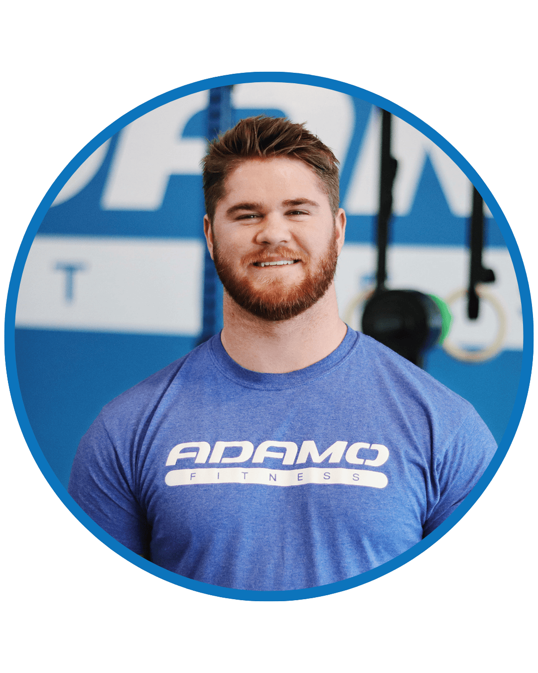 Smiling man with brown hair and beard wearing a blue Adamo Fitness t-shirt in a gym setting.