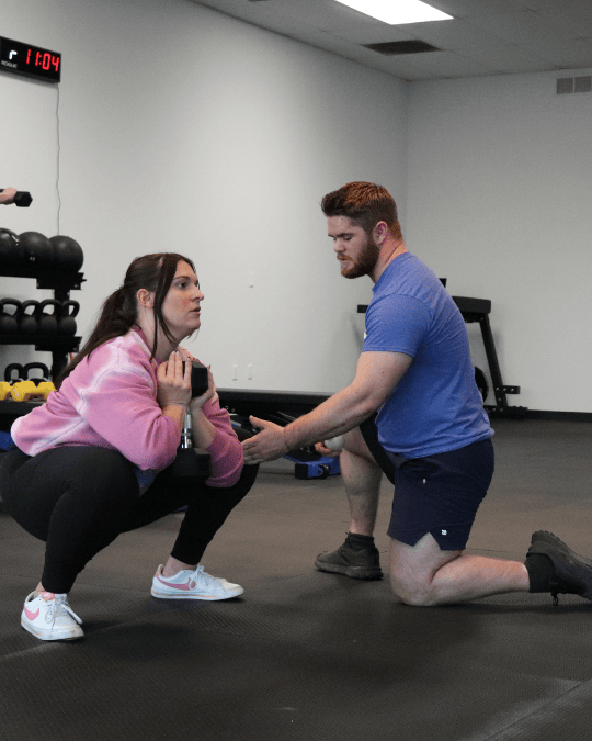 A woman in pink hoodie squats holding a dumbbell while a personal trainer kneels and coaches her in a gym.