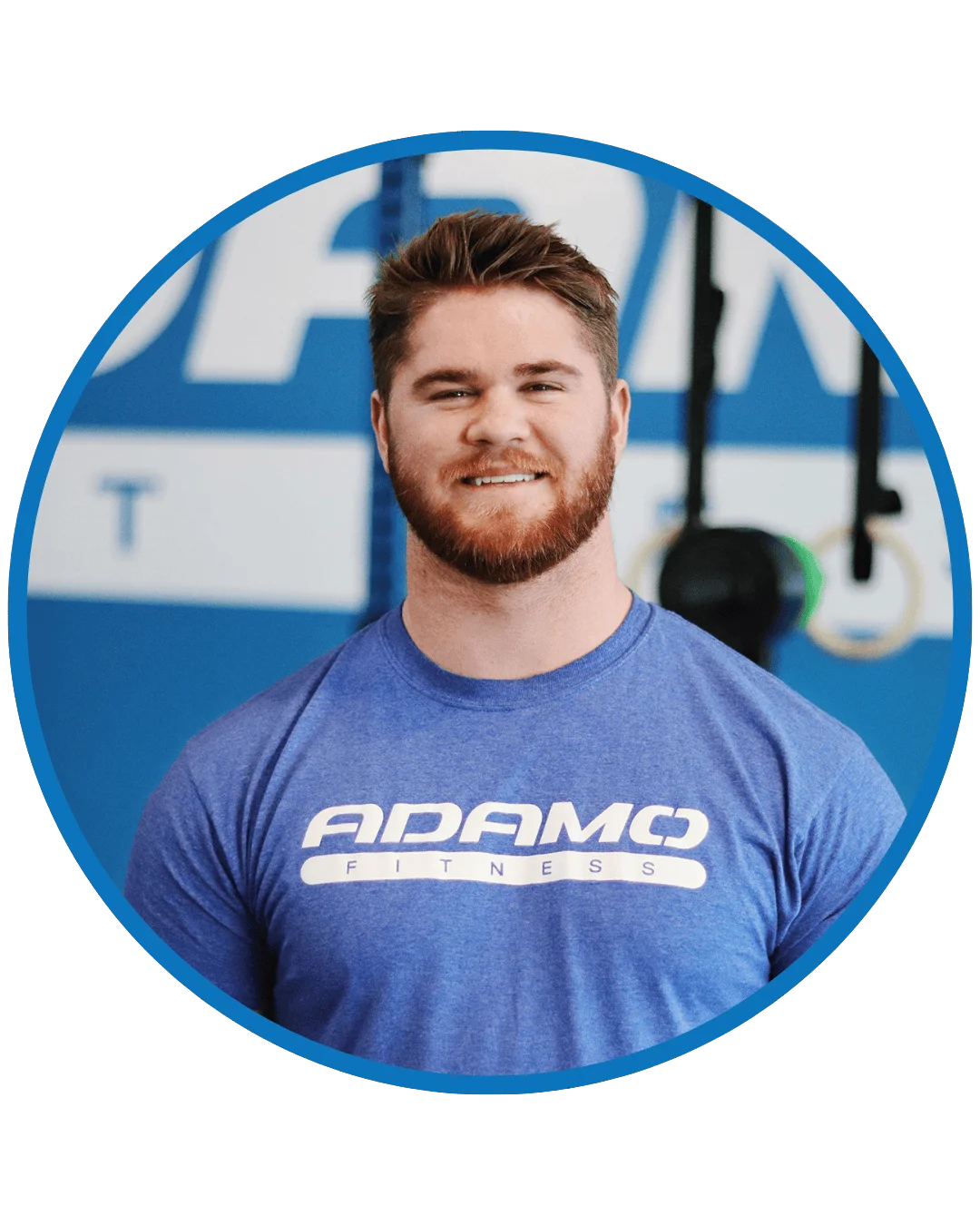 Smiling man with brown hair and beard wearing a blue Adamo Fitness t-shirt in a gym setting.