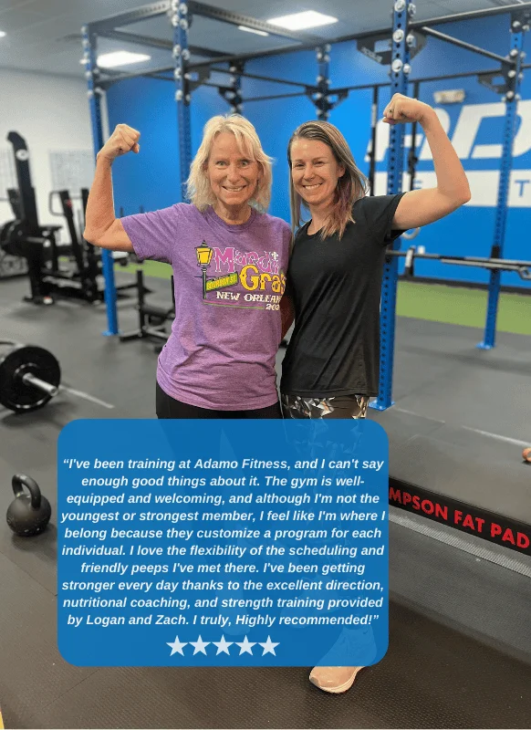 Two women smiling and flexing their arm muscles inside a gym with fitness equipment in the background.