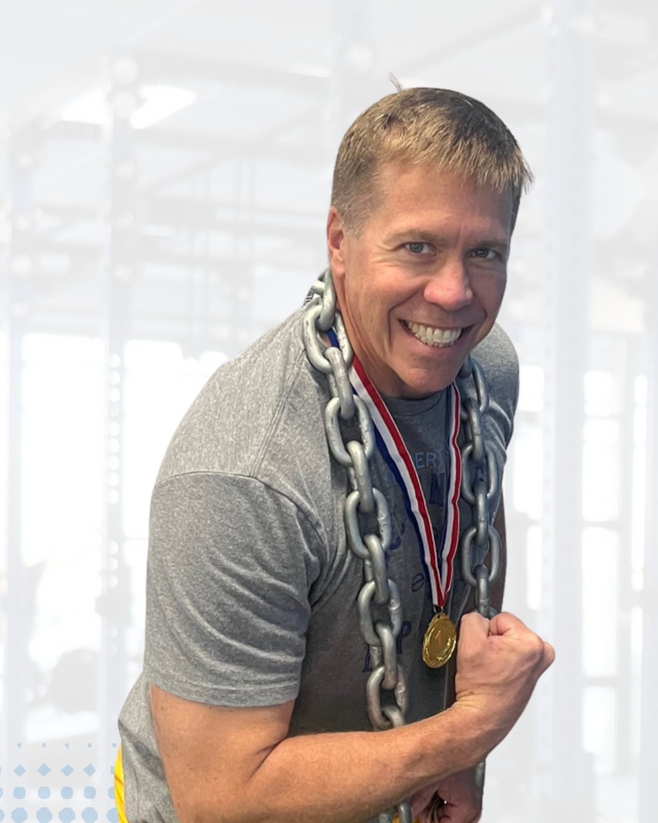 Tom, in his 50s, flexing with a lifting chain and medal at Adamo Fitness