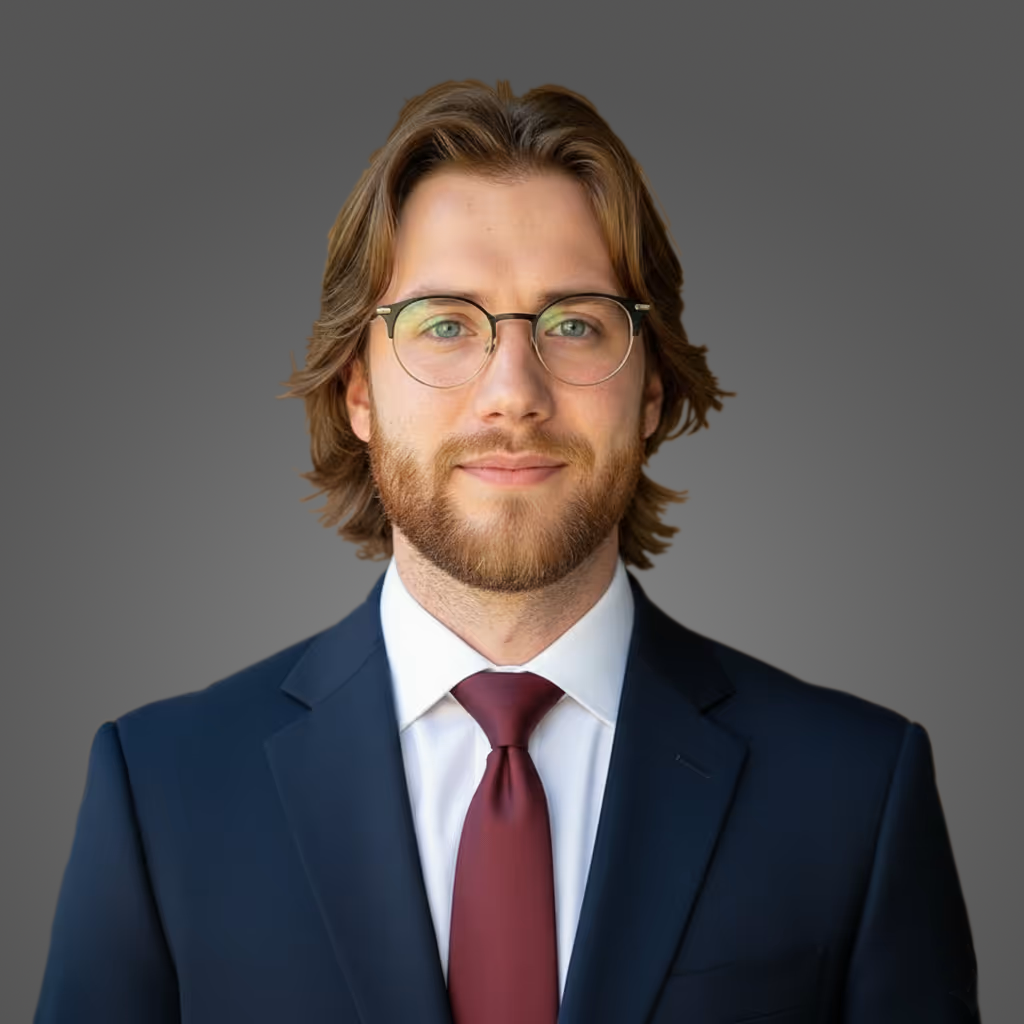 Portrait of a young man with glasses, medium-length light brown hair and beard, wearing a navy suit, white shirt, and maroon tie, against a gray background.