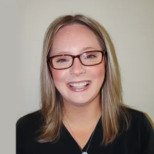 Smiling woman with blonde hair and glasses wearing a black top against a plain background.