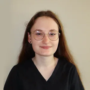 Young woman with long brown hair, glasses, and black top smiling against a plain background.