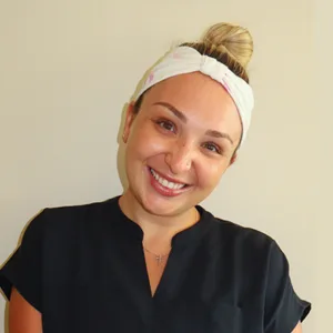 Smiling woman with a light headband and hair in a bun wearing a black top against a plain background.