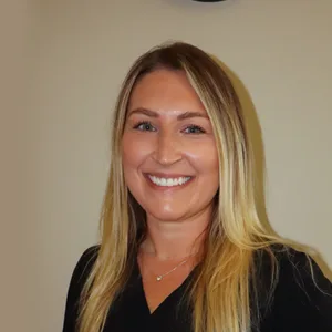 Smiling woman with long blonde hair wearing a black top against a beige background.
