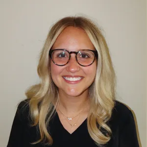 Smiling woman with blonde hair, wearing round eyeglasses and a black top against a plain background.