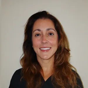 Smiling woman with long brown hair wearing a black top against a plain beige background.