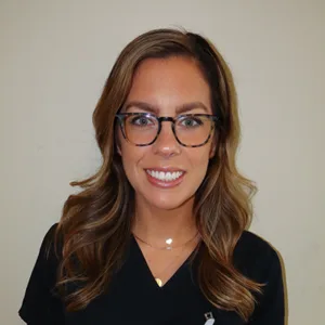 Smiling woman with long wavy brown hair and glasses wearing a black top against a plain background.