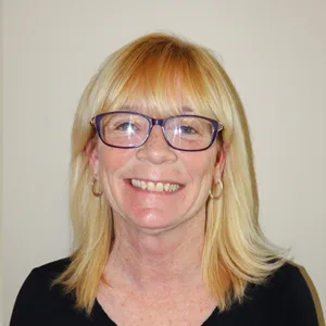 Smiling woman with straight blonde hair, glasses, and hoop earrings wearing a black top.