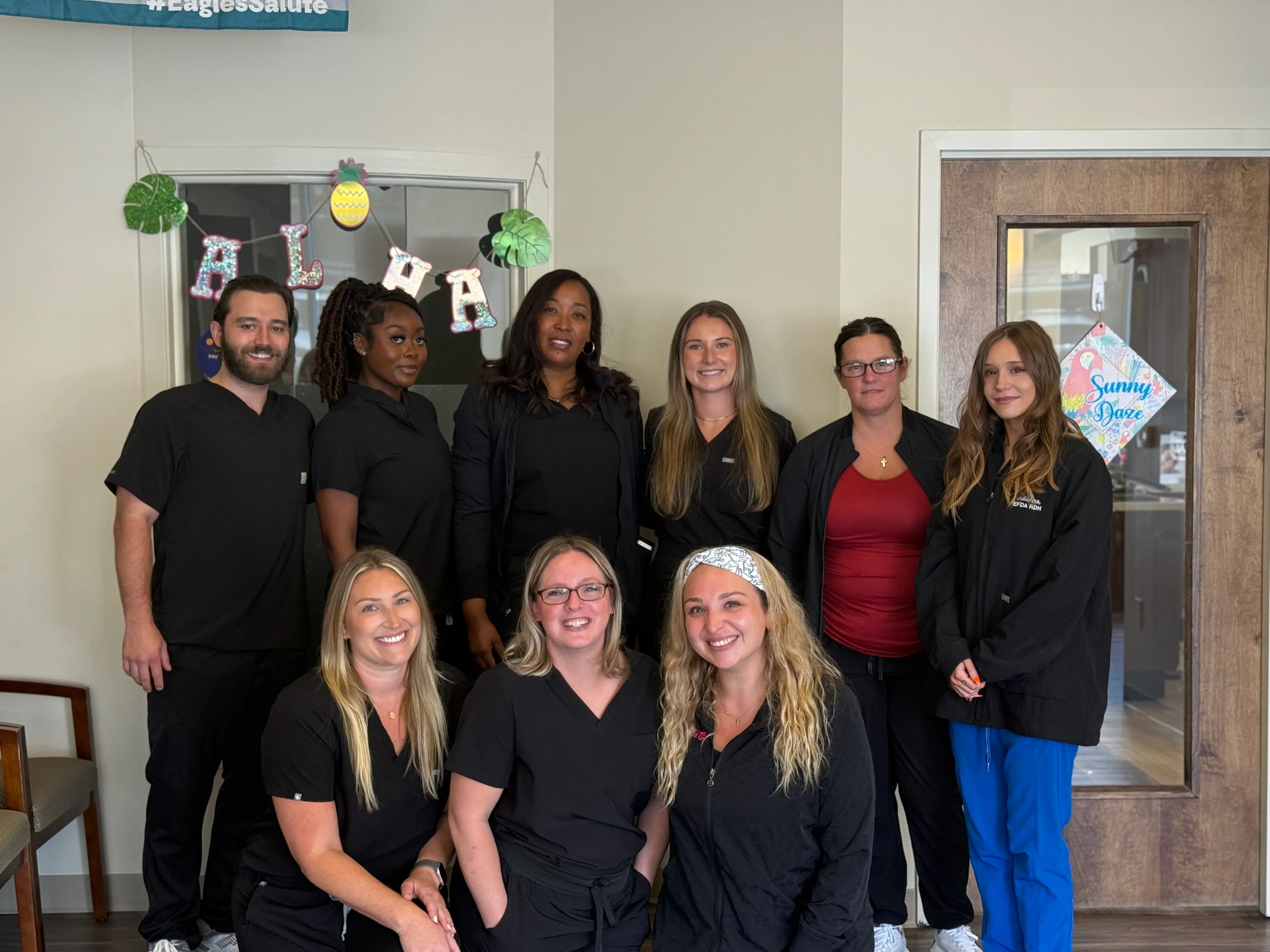 Group of nine people in black scrubs posing indoors under an 'ALOHA' decoration.