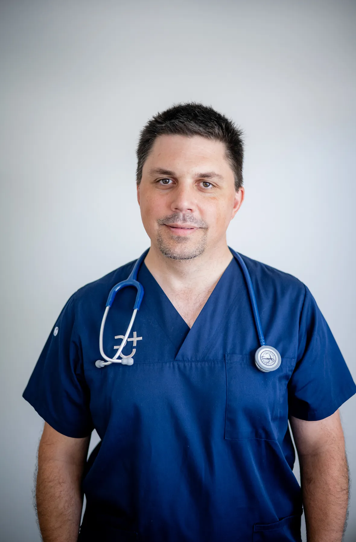 Porträt von Dr. Stefan Reschke in blauer medizinischer Kleidung mit einem Stethoskop um den Hals vor grauem Hintergrund.