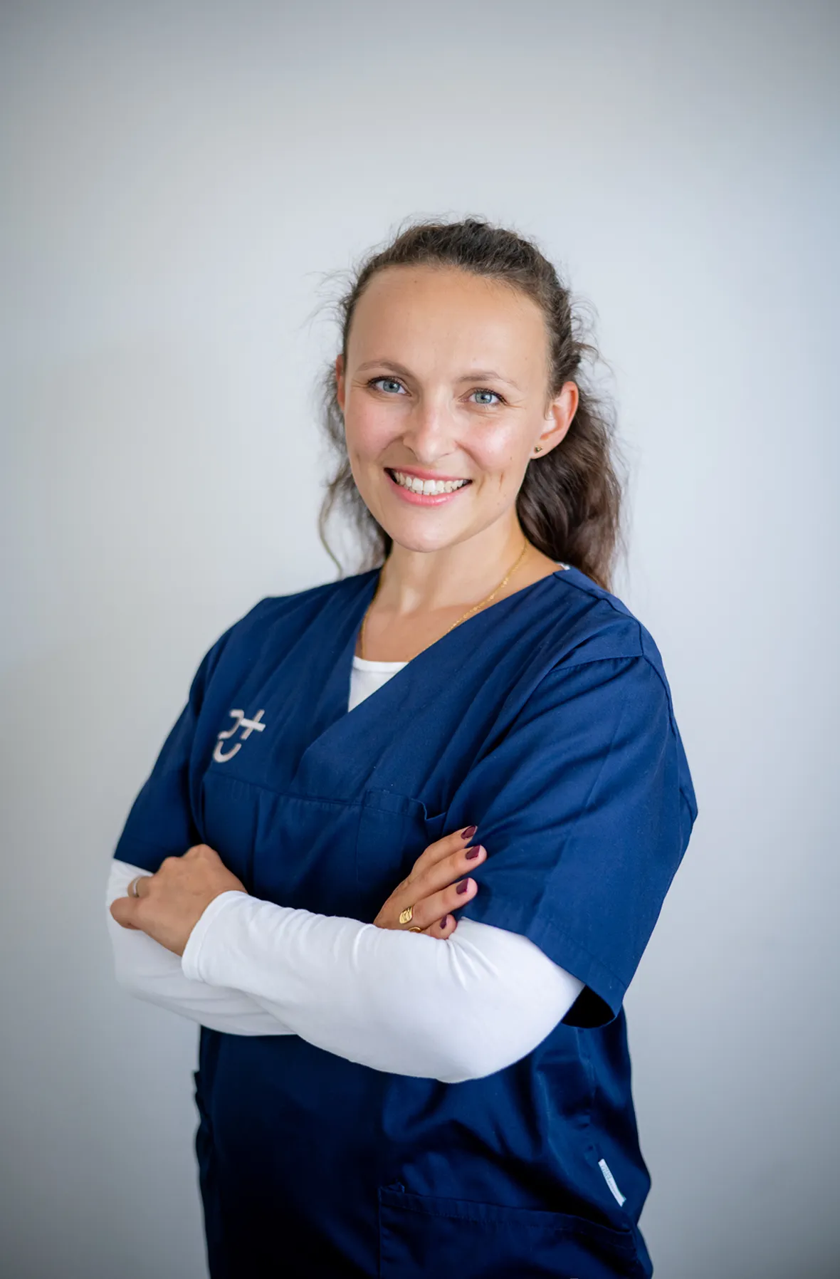 Lächelnde Natalia Schrank mit zusammengelegten Armen, trägt blaue medizinische Arbeitskleidung vor einem hellen Hintergrund.