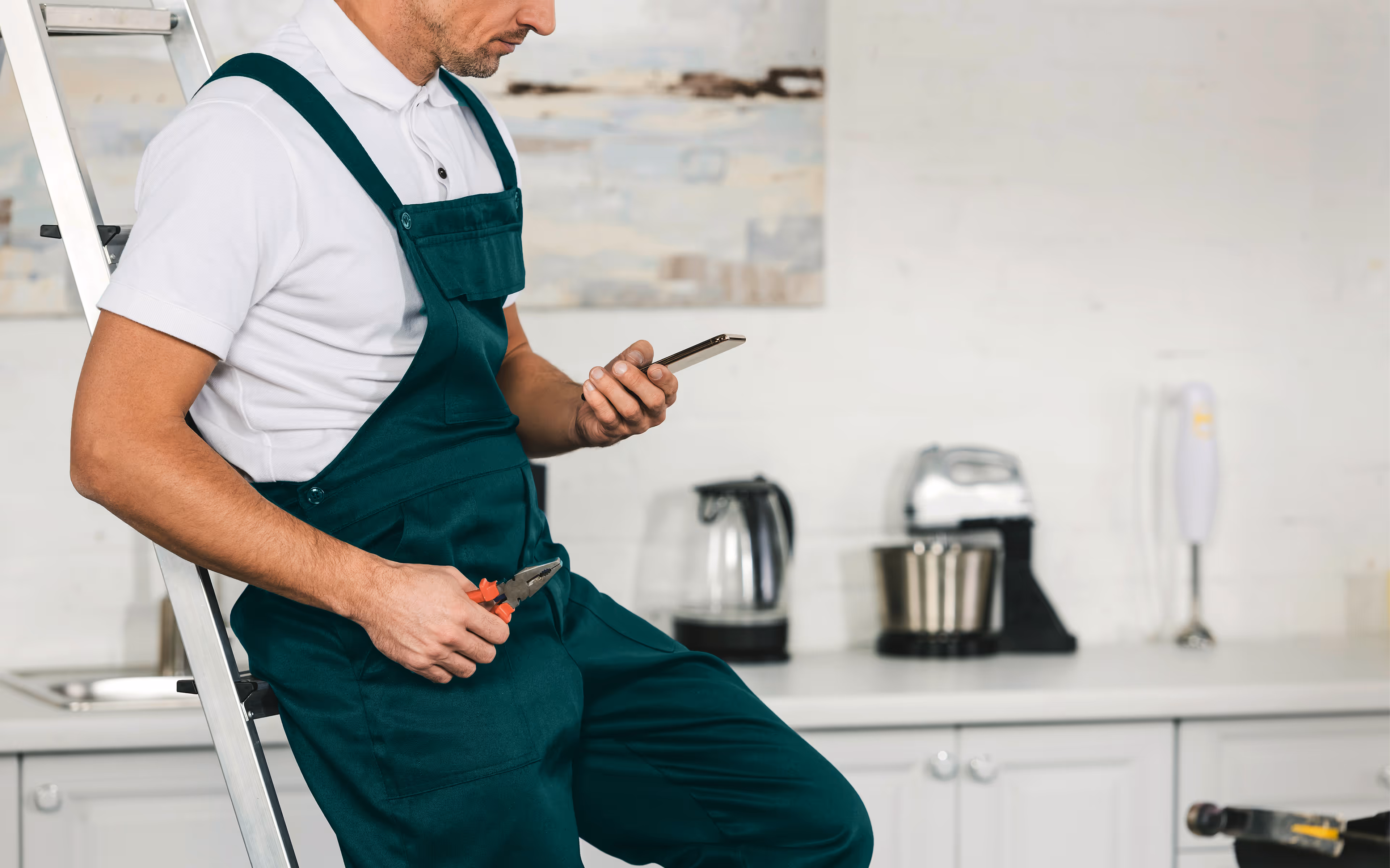 Handwerker in grünem Overall sitzt auf einer Leiter und benutzt ein Smartphone, hält eine Zange in der anderen Hand.