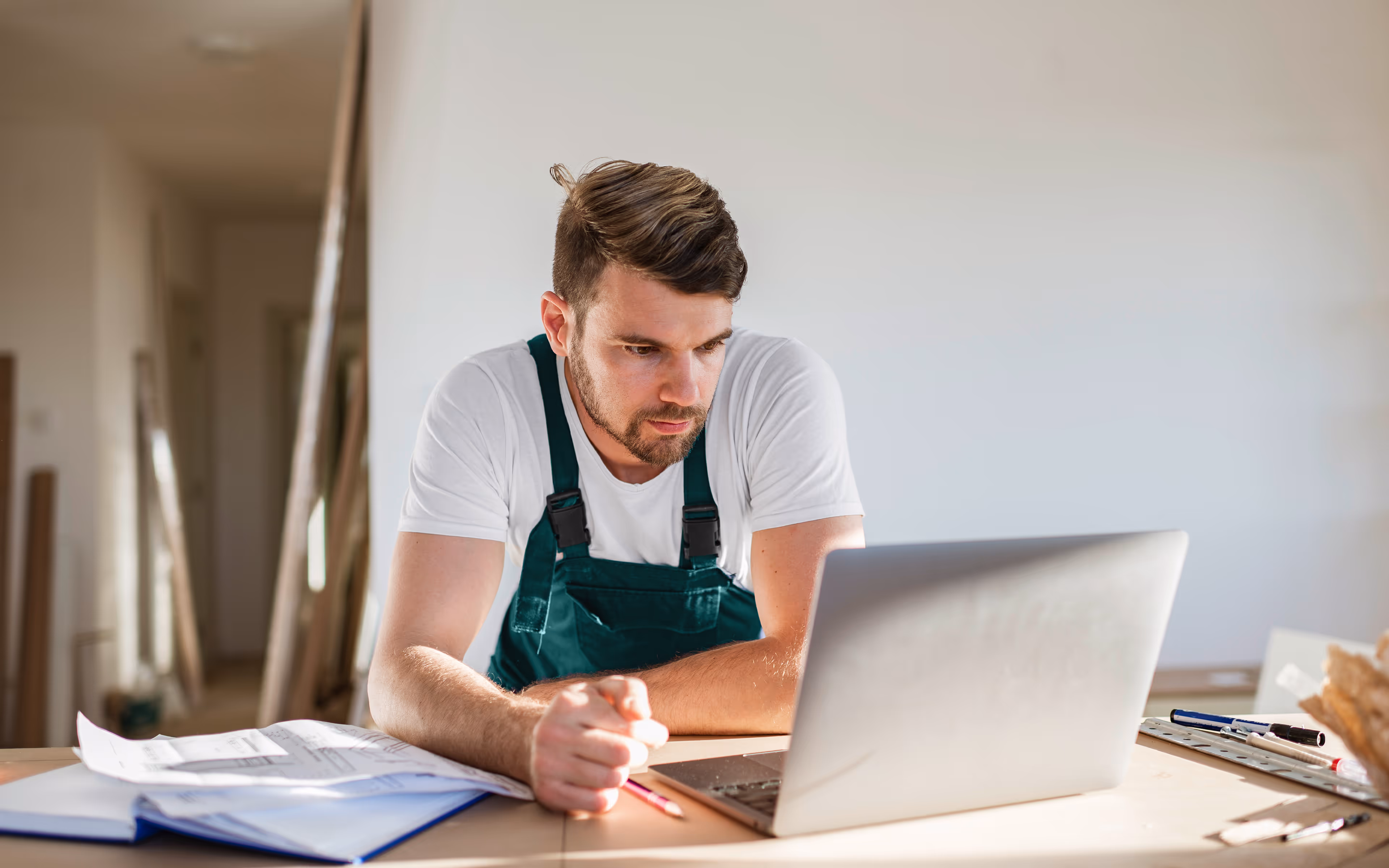 Handwerker in grüner Arbeitskleidung sitzt an einem Tisch und arbeitet konzentriert an einem Laptop mit Planungsunterlagen neben sich.