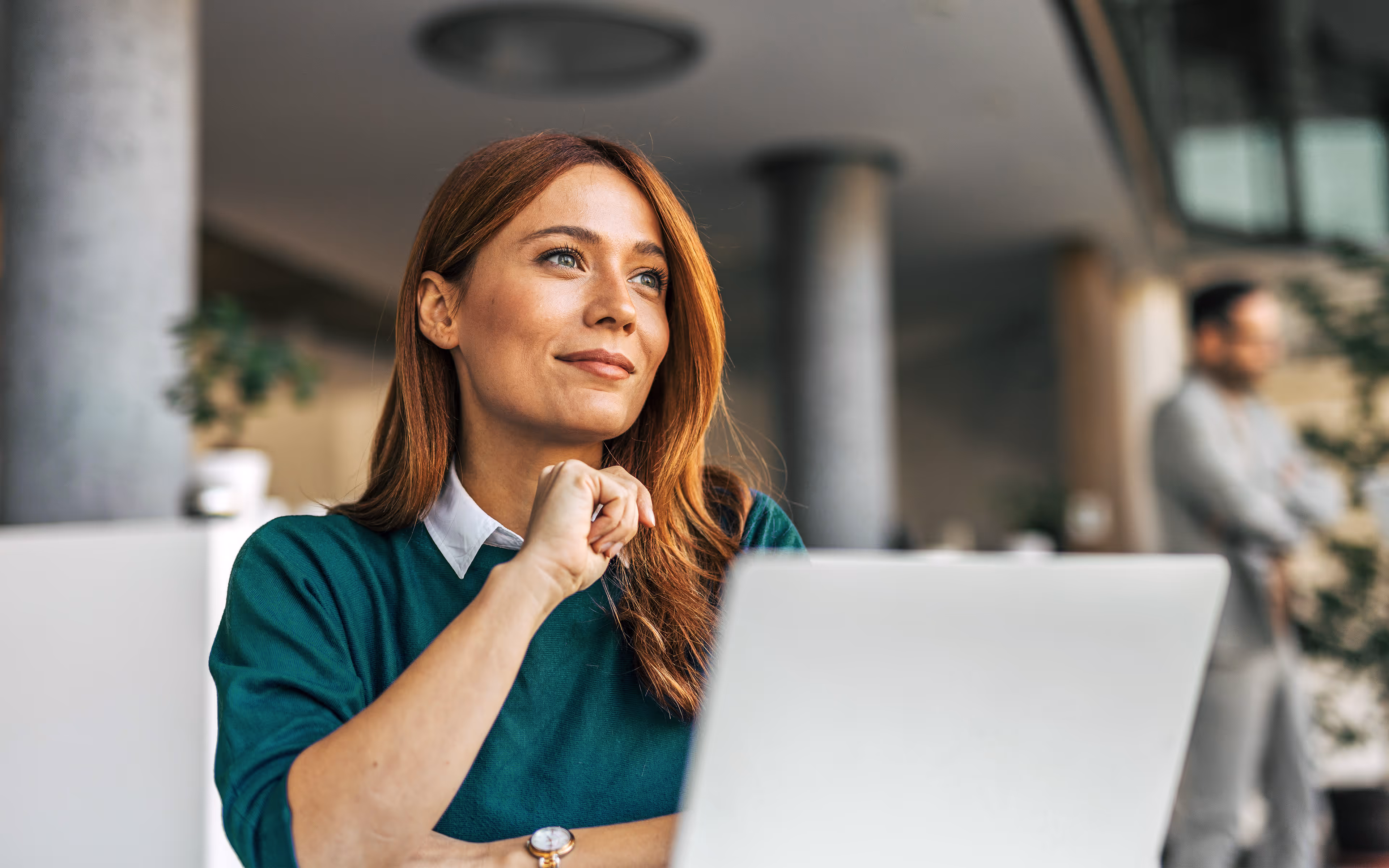 Zufriedene rothaarige Frau sitzt vor einem Laptop in einem modernen Büro und blickt entspannt in die Ferne.