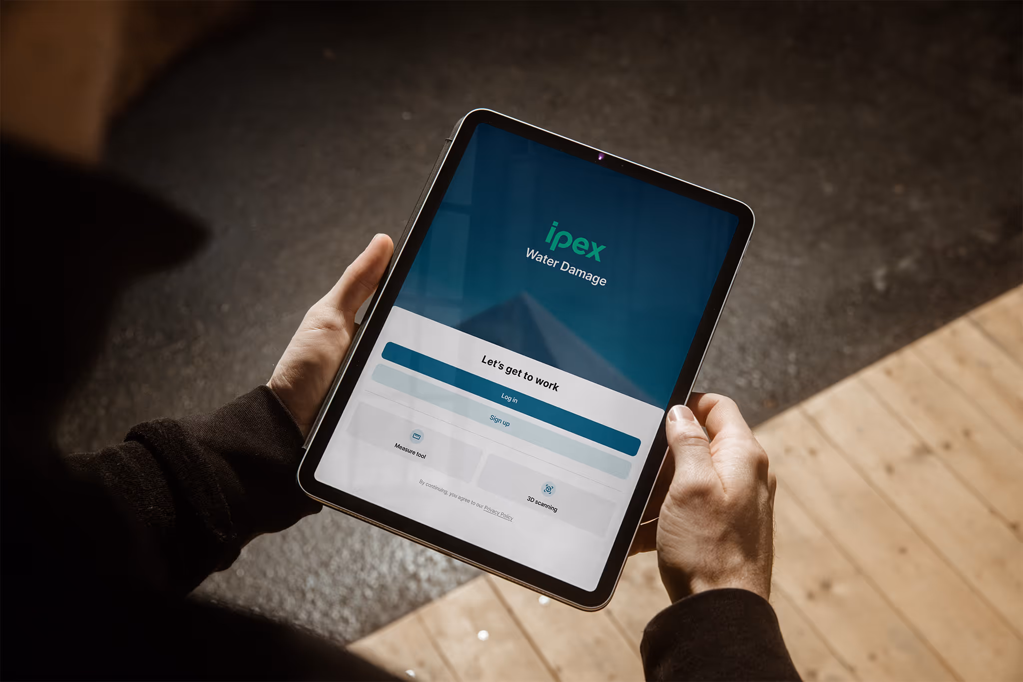 Person hält ein iPad mit der Ipex Wasserschaden-App geöffnet, Anmeldebildschirm mit Optionen für Login, Registrierung, Messwerkzeug und 3D-Scan.