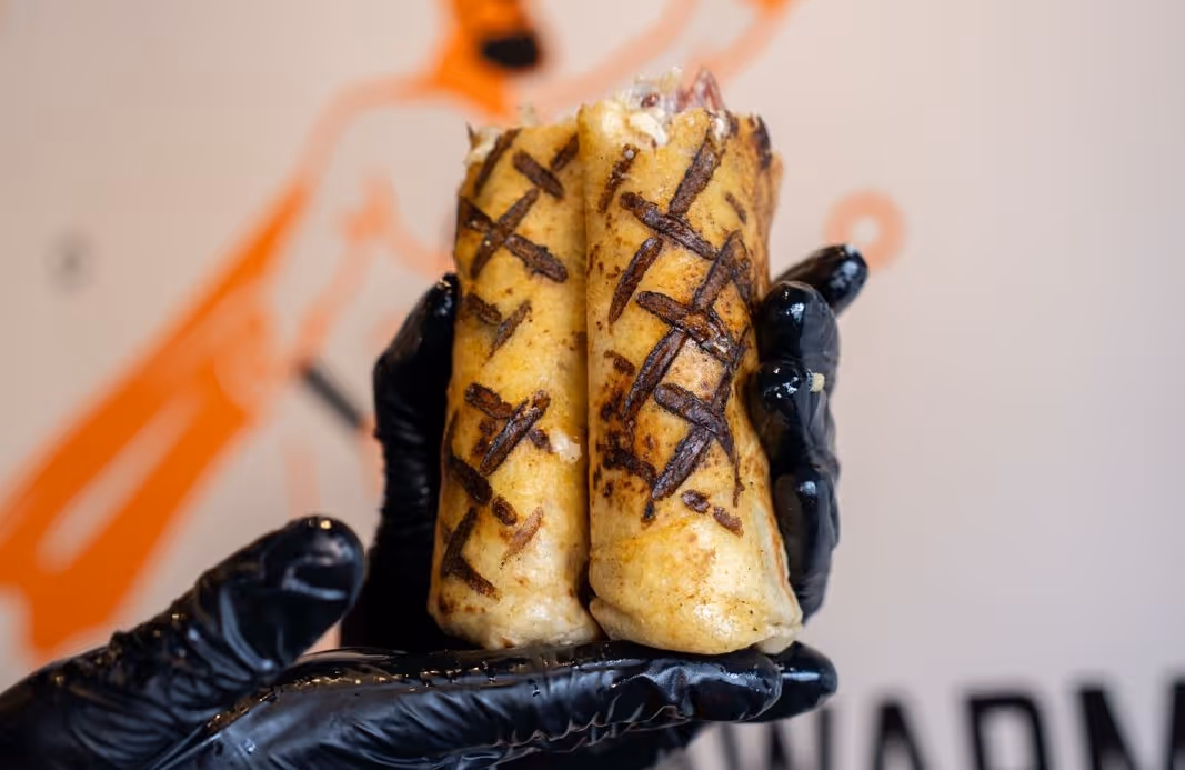 Two grilled wraps held together by gloved hands with a blurred orange and white background.