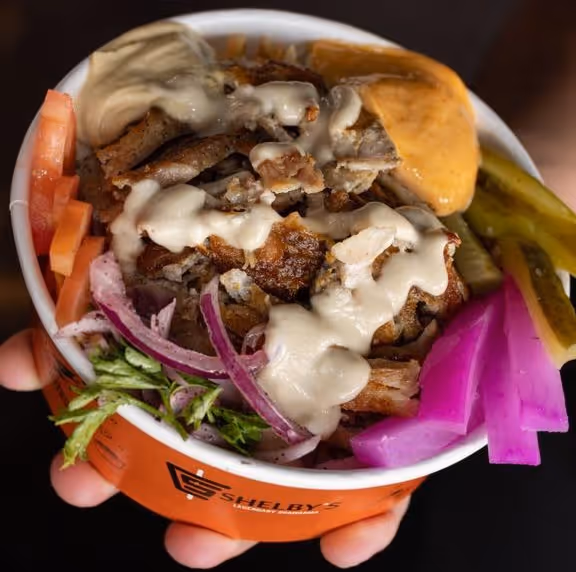 Hand holding a bowl of grilled meat topped with creamy sauce, surrounded by sliced tomatoes, onions, pickles, and purple pickled vegetables.