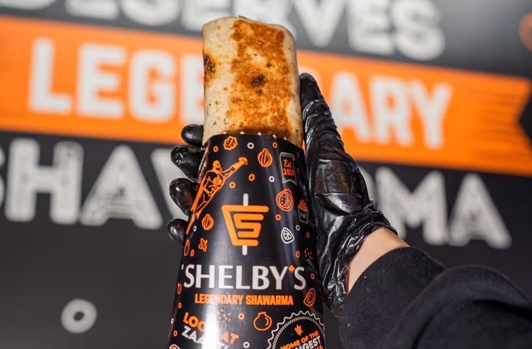 Hand wearing black glove holding a wrapped, grilled shawarma labeled 'Shelby's Legendary Shawarma' with blurred background text.